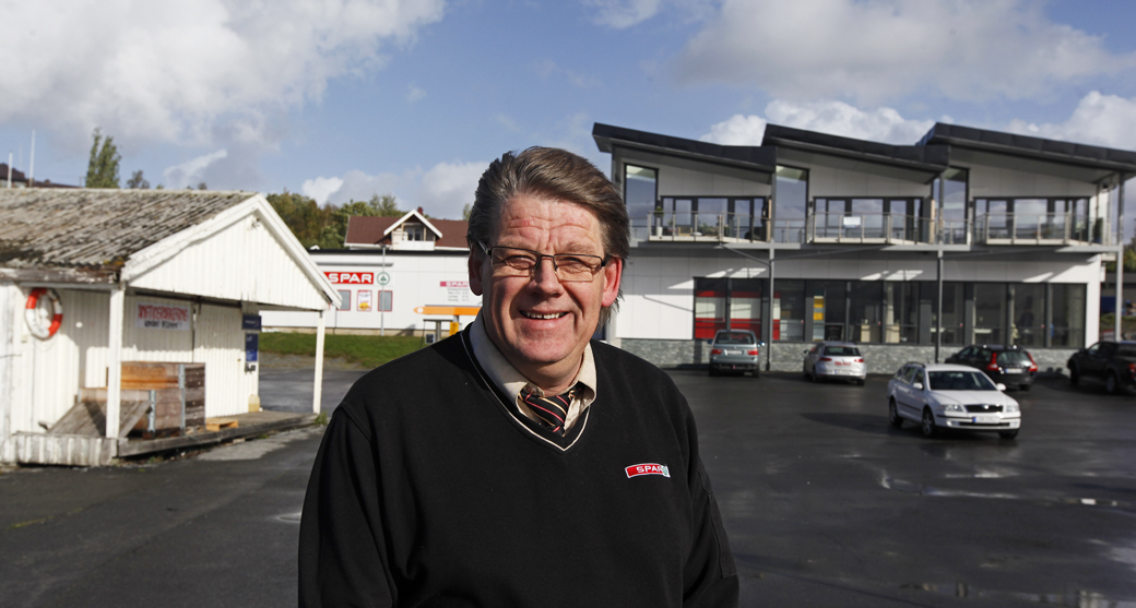 Johan Sørgaard foran den gamle og nye tid. Melbrygga til venstre ble bygd i 1920 og berget unna brannen i 1954. Nybutikken med leiligheter i andre etasje sto ferdig i 2012.